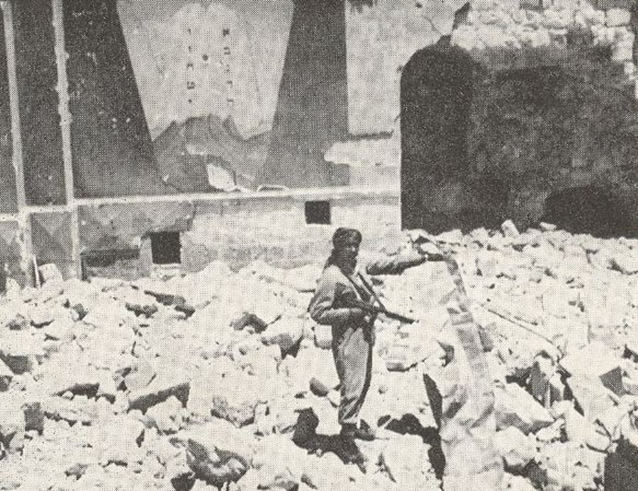 חייל הלגיון הערבי ניצב בביהכ"נ החורבה בירושלים, ומחזיק בידו ספר תורה מחולל, 1948
