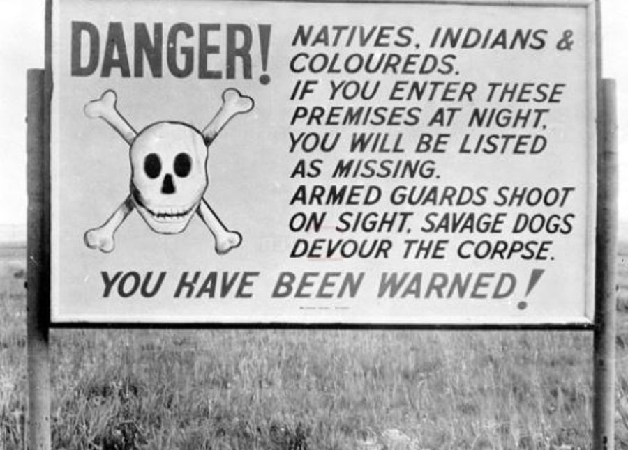 שלט: 'סכנה! לאומיים, אינדיאנים וצבעוניים, אם תיכנסו לשטח זה בלילה, תירשמו כנעדרים. שומרים חמושים יורים במקום, כלבים פראיים זוללים את הגופות. ראו הוזהרתם!'. יוהנסבורג, דרום אפריקה, 1953