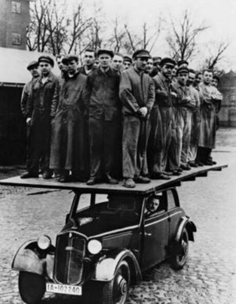 עשרות אנשים עומדים על לוח שהוצב על גבי מכונית קטנה, כדי להוכיח את חוזקה. שנות ה-1930